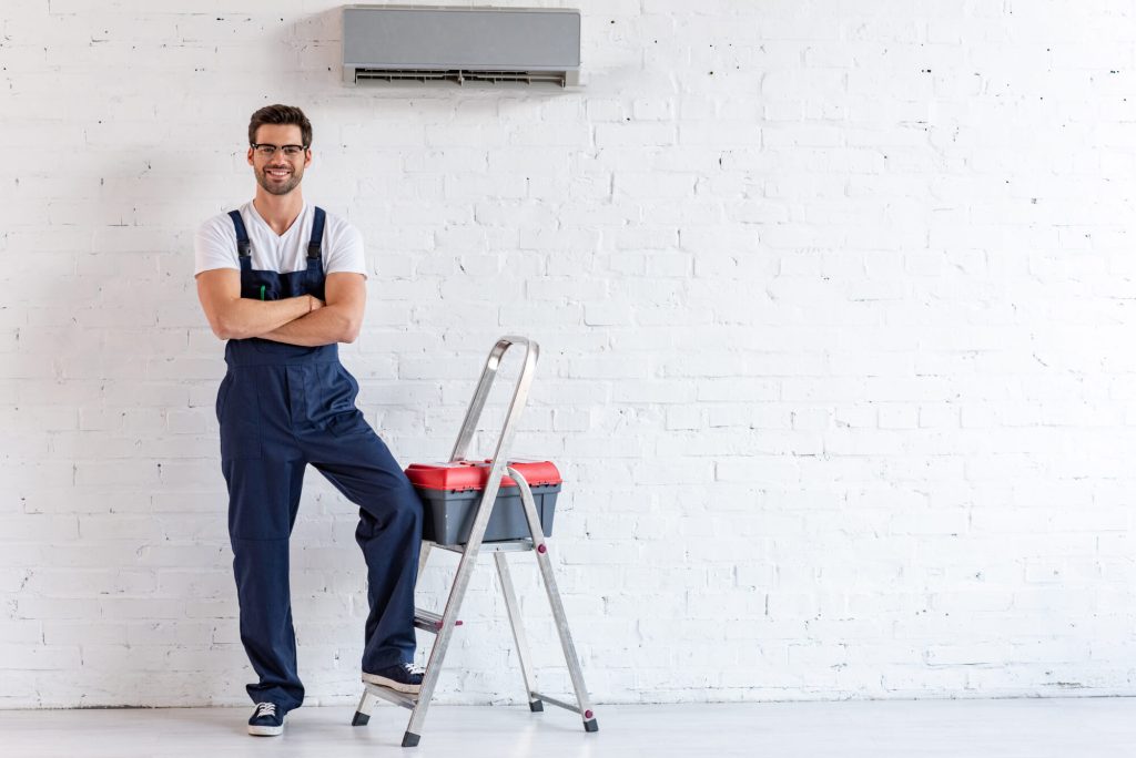 A man in overalls stands beside a wall-mounted air conditioner, showcasing a casual and practical setting.